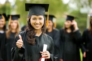 A woman with a medium complexion and brown hair stands in front of her peers, all wearing caps and gowns, and smiles while holding a diploma and giving a thumbs up.