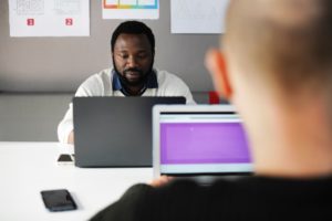 A black man works at his laptop opposite his white coworker at a shared work table with print materials hanging on a presentation board behind him.
