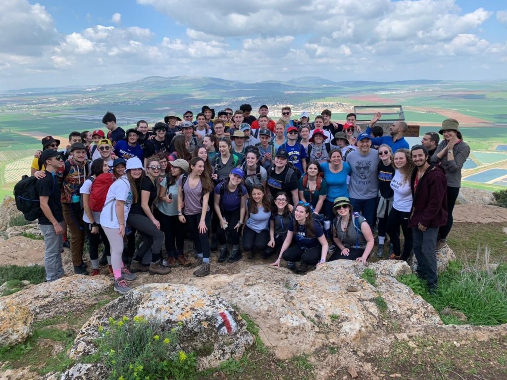 A large group of 82 students from two Jewish day schools in Washington, D.C. are smiling and posing for a photo at the top of Mt. Gilboa in Israel.