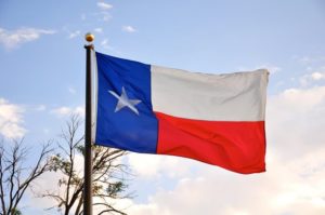 Flag of Texas flying on a pole against a bright sky with some clouds and treetops.