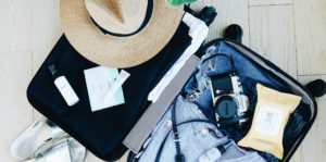 A hard-shell spinner carry-on sits open on cream colored flooring. Denim shorts, striped floral top, sun hat, lotion, face wipes, and a Nikon film camera sit inside and a pair of silver sandals sits next to the luggage.