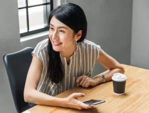 A Japanese woman sits at a table with her coffee holding her phone and looking to her right while smiling.