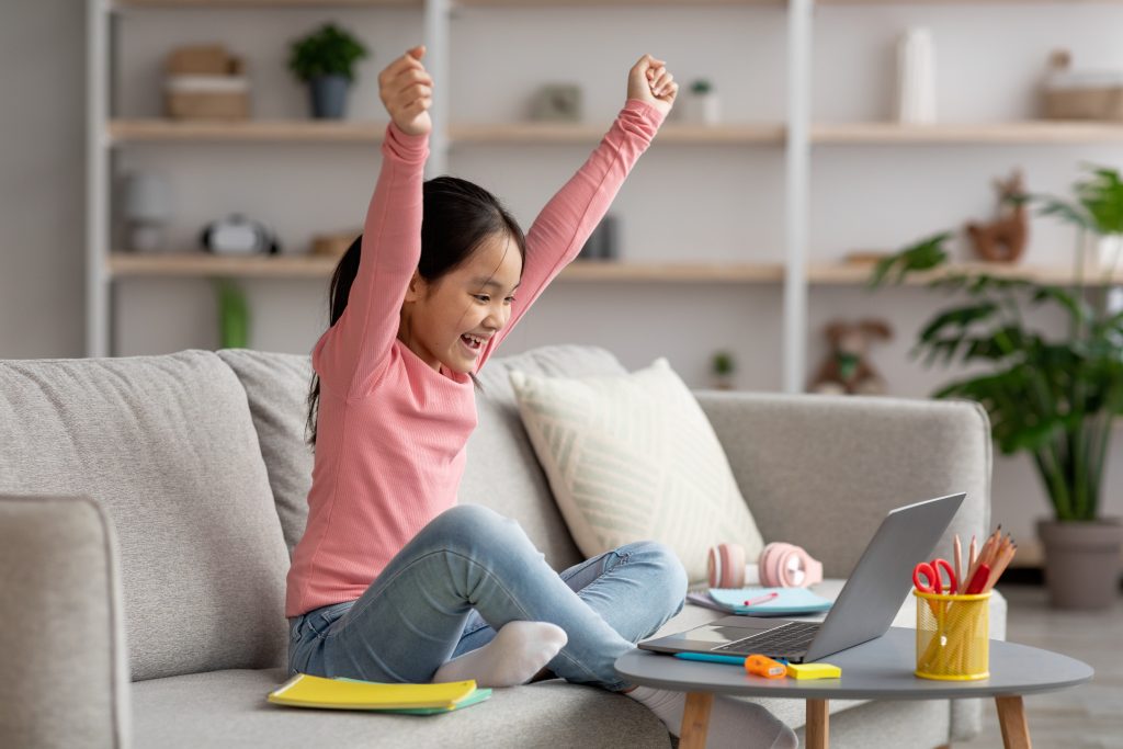 Young girl looking at her computer screen and raising her hands in success